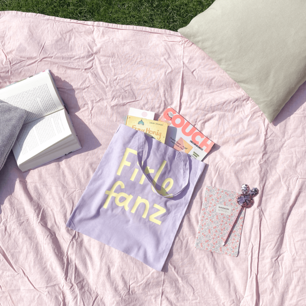 Stofftasche in lila liegt auf einer Picknickdecke mit ein paar Büchern un der Zeitschrift Couch